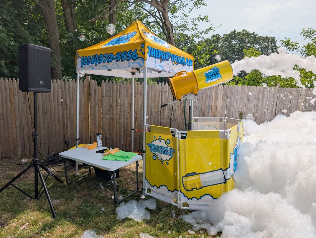 A foam cannon blowing a pile of foam across a backyard, with the tent and foam pit walls set up on the grass